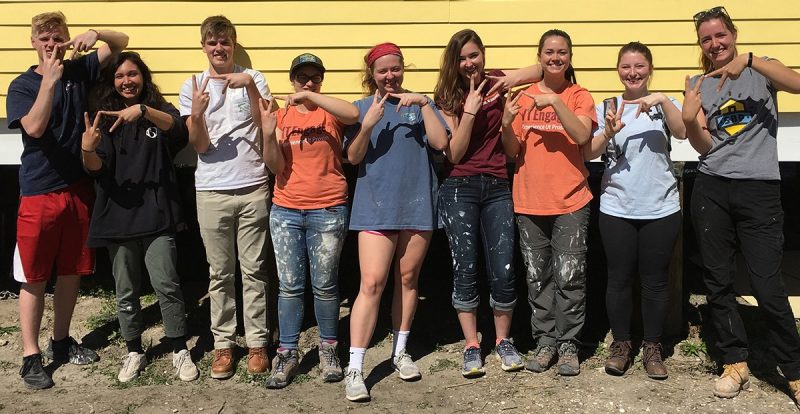 A group of students pose in front of a yellow house and make the "VT" sign with their hands.